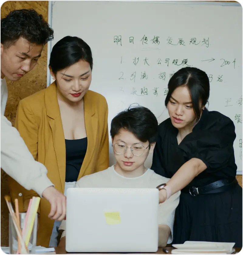 Team collaborating around a laptop, with a whiteboard in the background displaying notes.