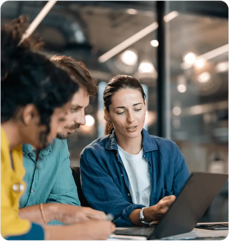 Three colleagues discussing work, with one presenting ideas on a laptop in a modern office setting.