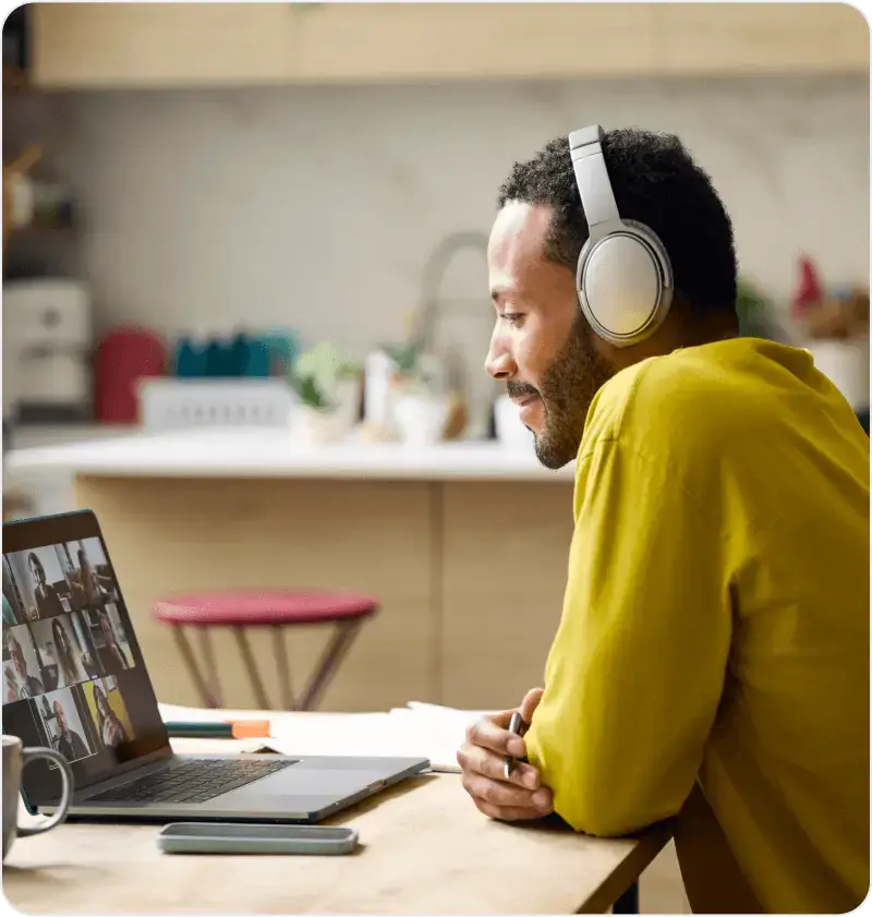 A man in headphones participates in a video call on a laptop while sitting in a modern kitchen.