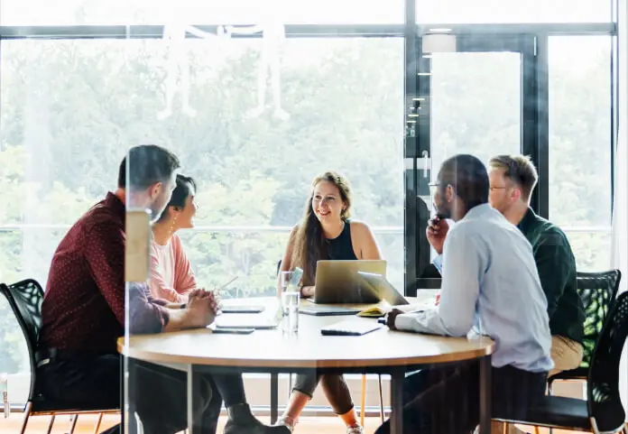 A group of colleagues having a meeting around a round table in a bright, glass-walled room.