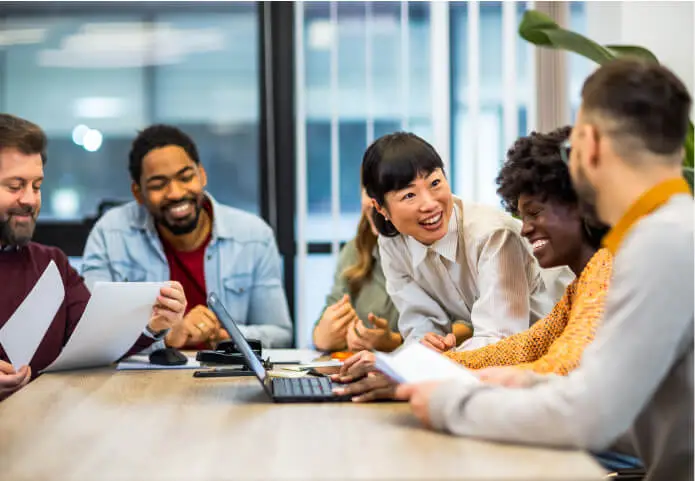 A diverse group of coworkers smiling and collaborating around a table with documents and a laptop.